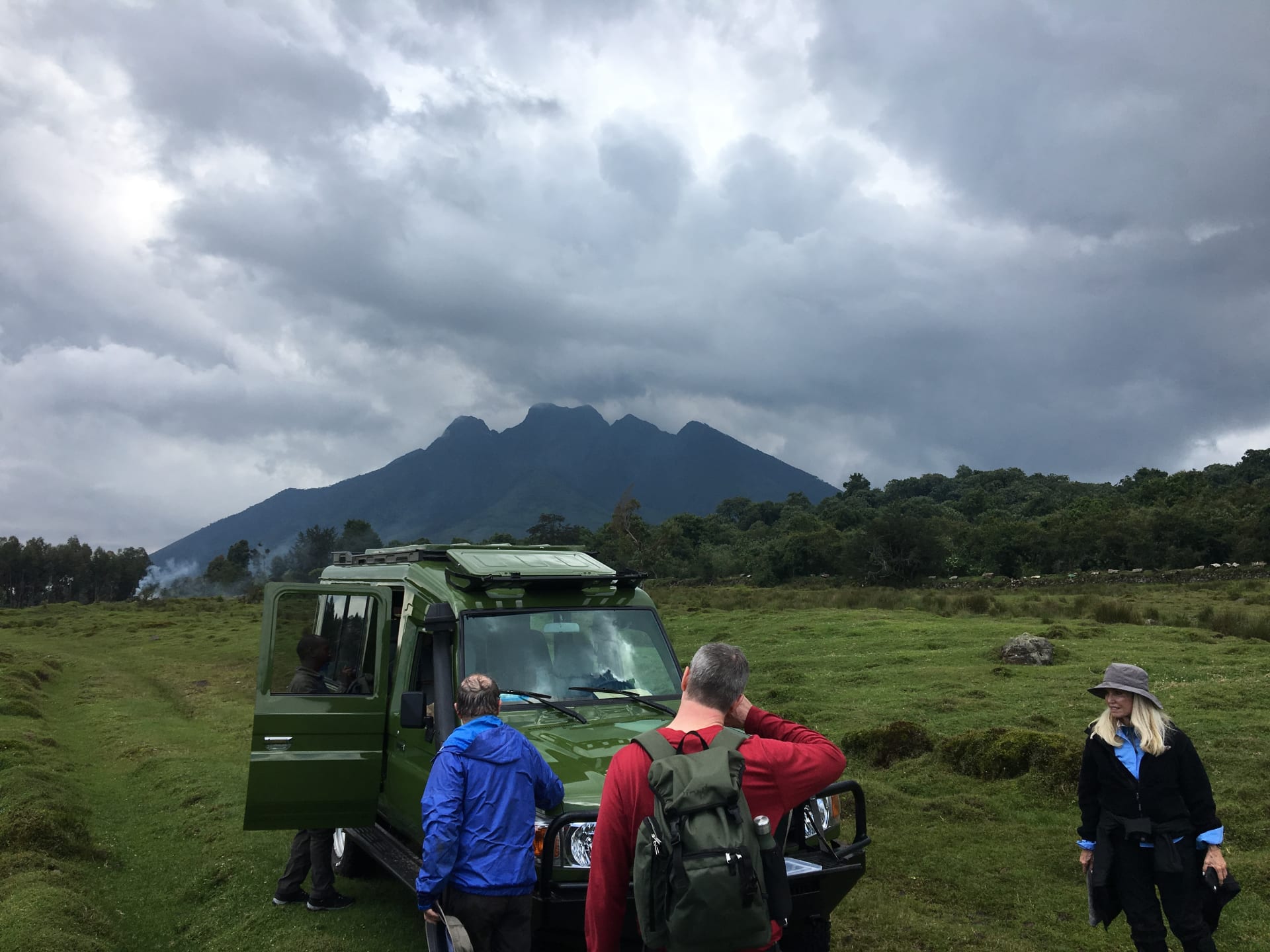 Travellers beside a safari vehicle with a volcano behind them near Volcanoes National Park, Rwanda