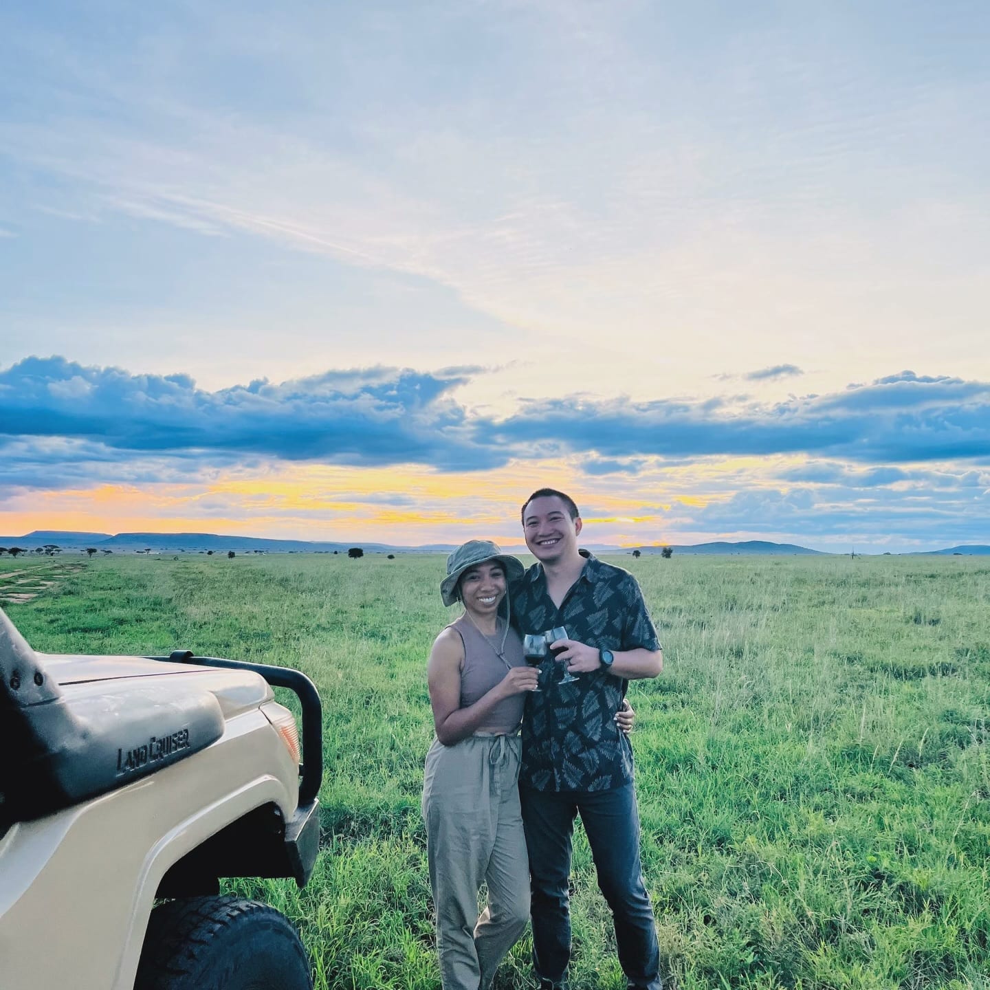 A couple standing on green plains beside a safari vehicle under a colourful sky
