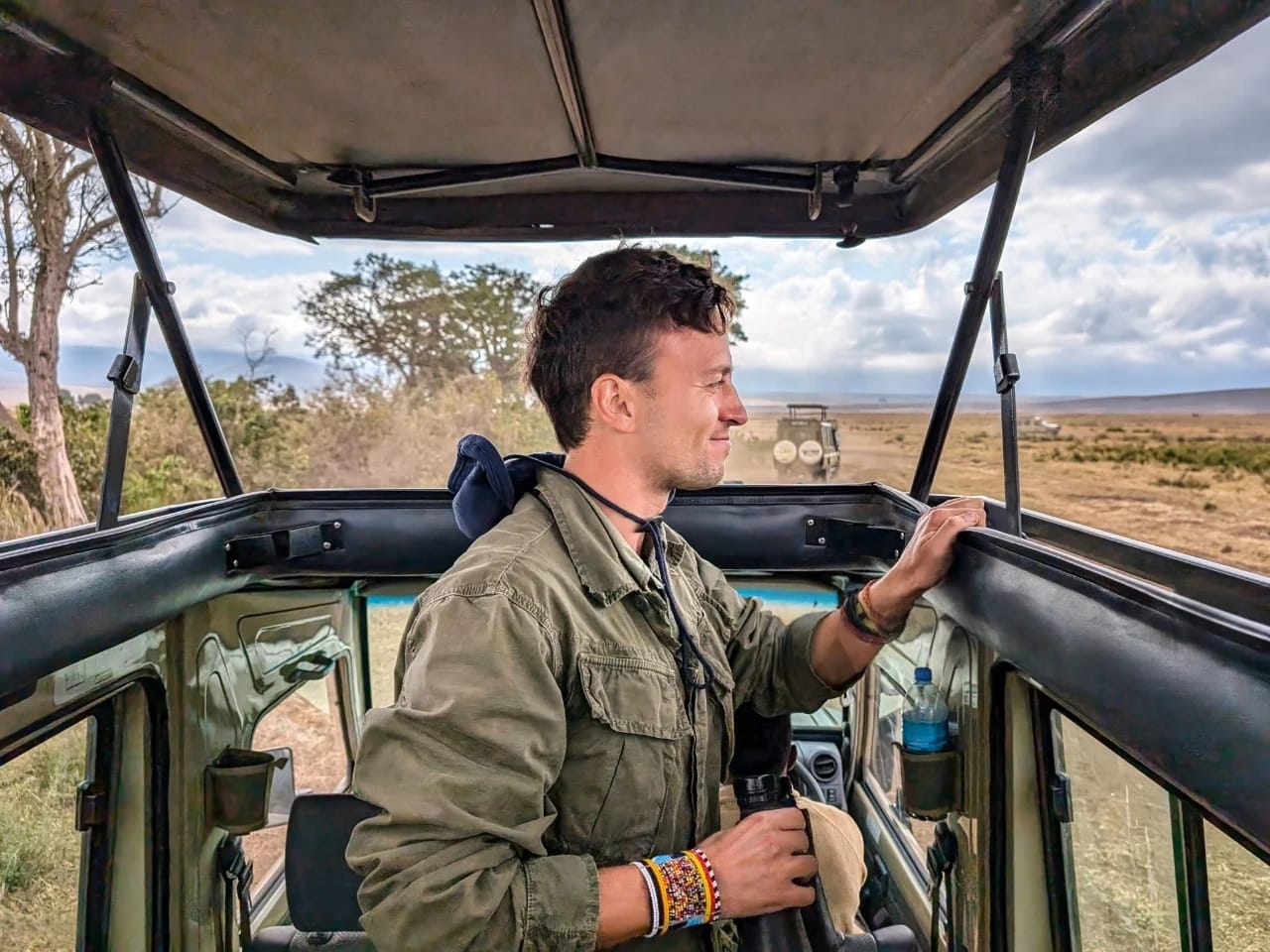 Man standing at the open roof of a safari vehicle in Serengeti, Tanzania, watching wildlife and vast golden plains — perfect visual for a guide on what to pack for Tanzania safari.