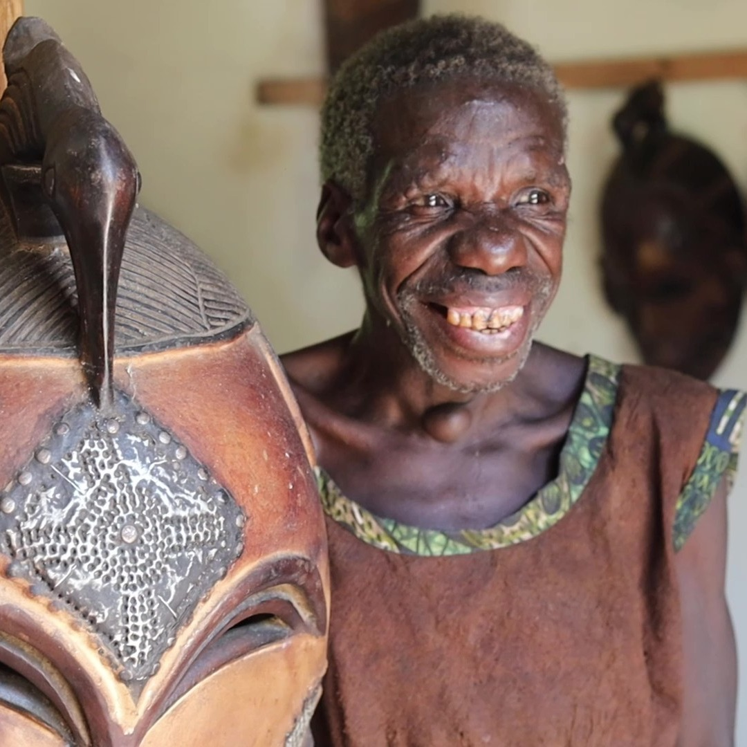 An elderly Batwa woman beside a carved mask during a cultural visit