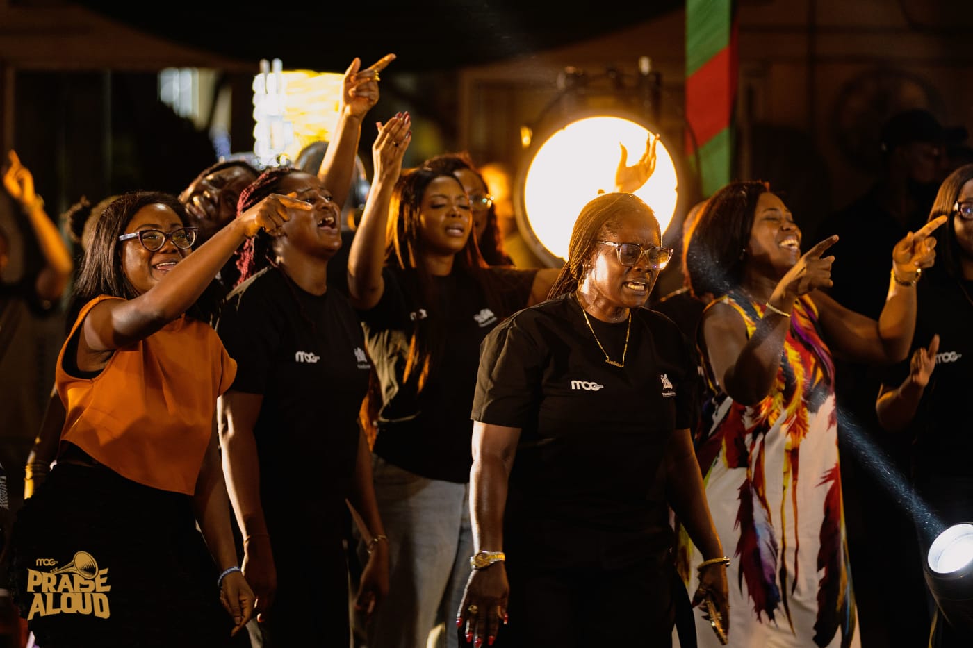 Worshippers at MOG Praise Aloud singing and pointing upward, lit by warm stage lights