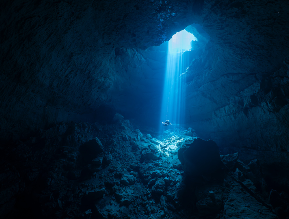Diver in underwater cave with a light shining on them.