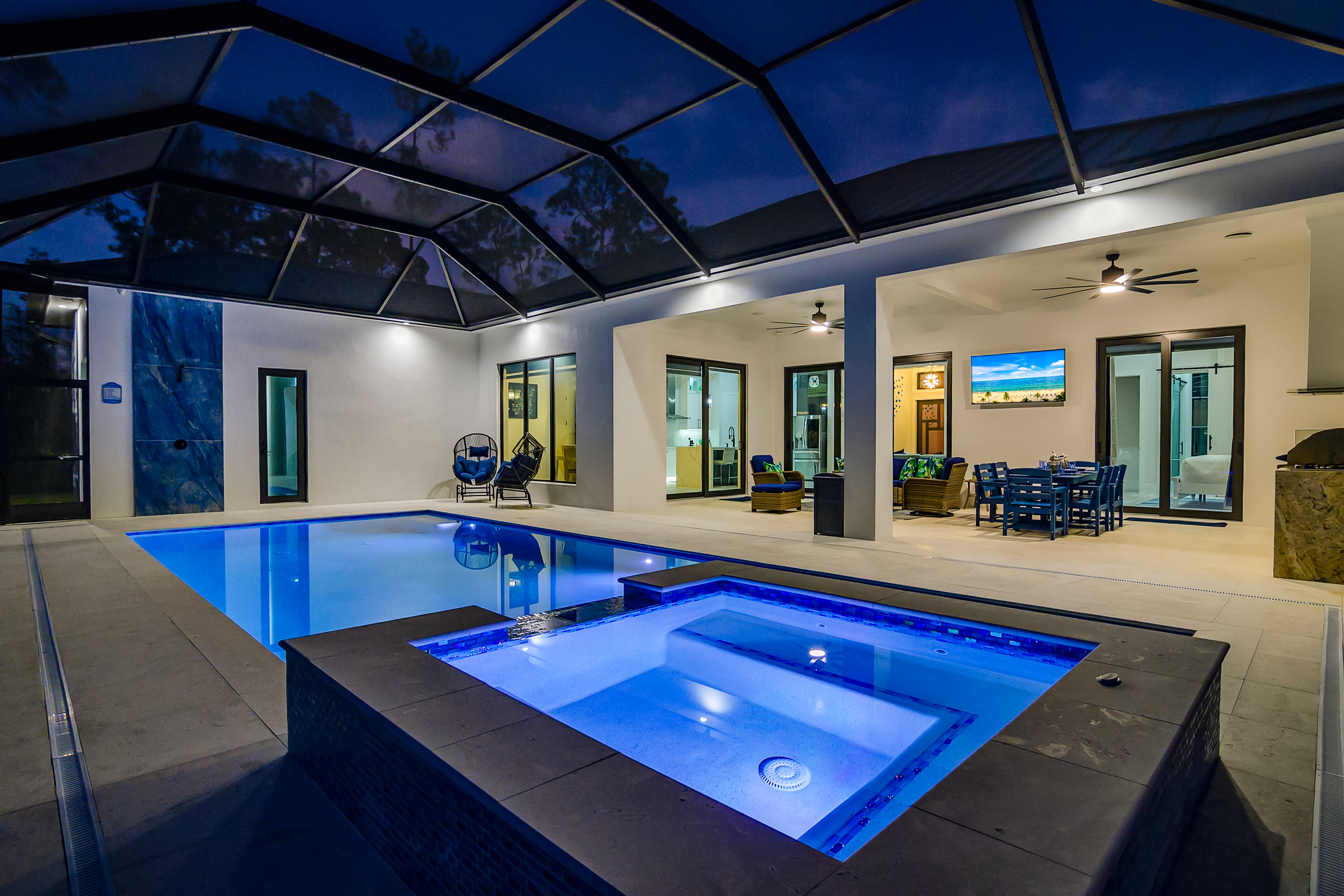 Swimming pool and spa with blue lighting under screened enclosure at night showing seating and kitchen area