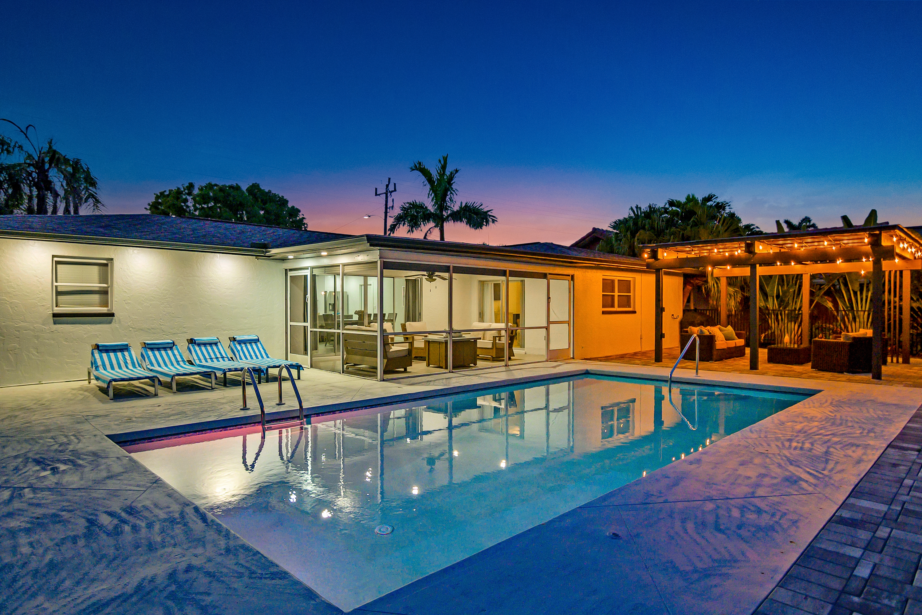 Swimming pool at night with striped lounge chairs on left and lit pergola with seating on right