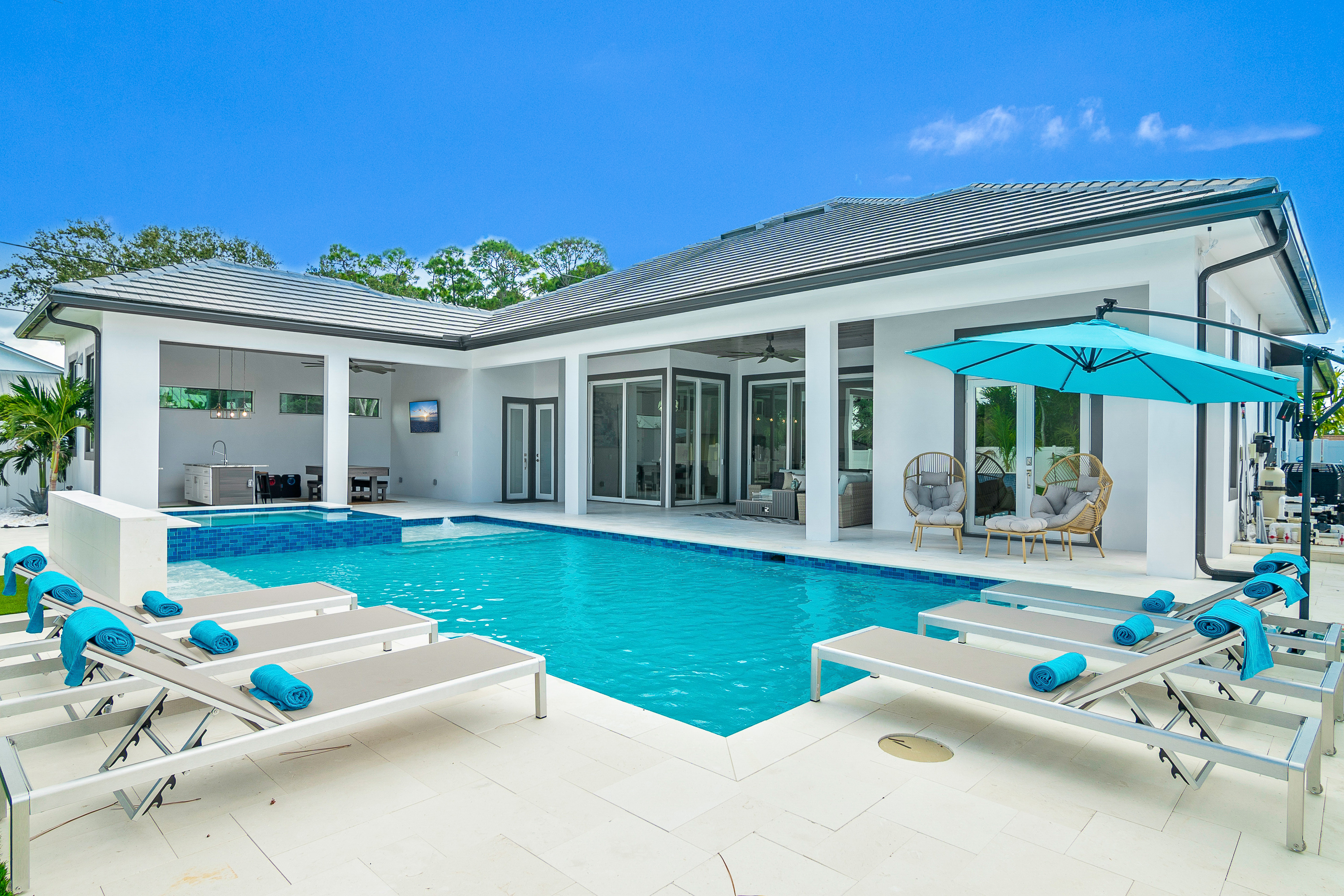 Modern outdoor pool with tiled spa area, white loungers with blue towels, and turquoise umbrella near white house exterior under clear sky