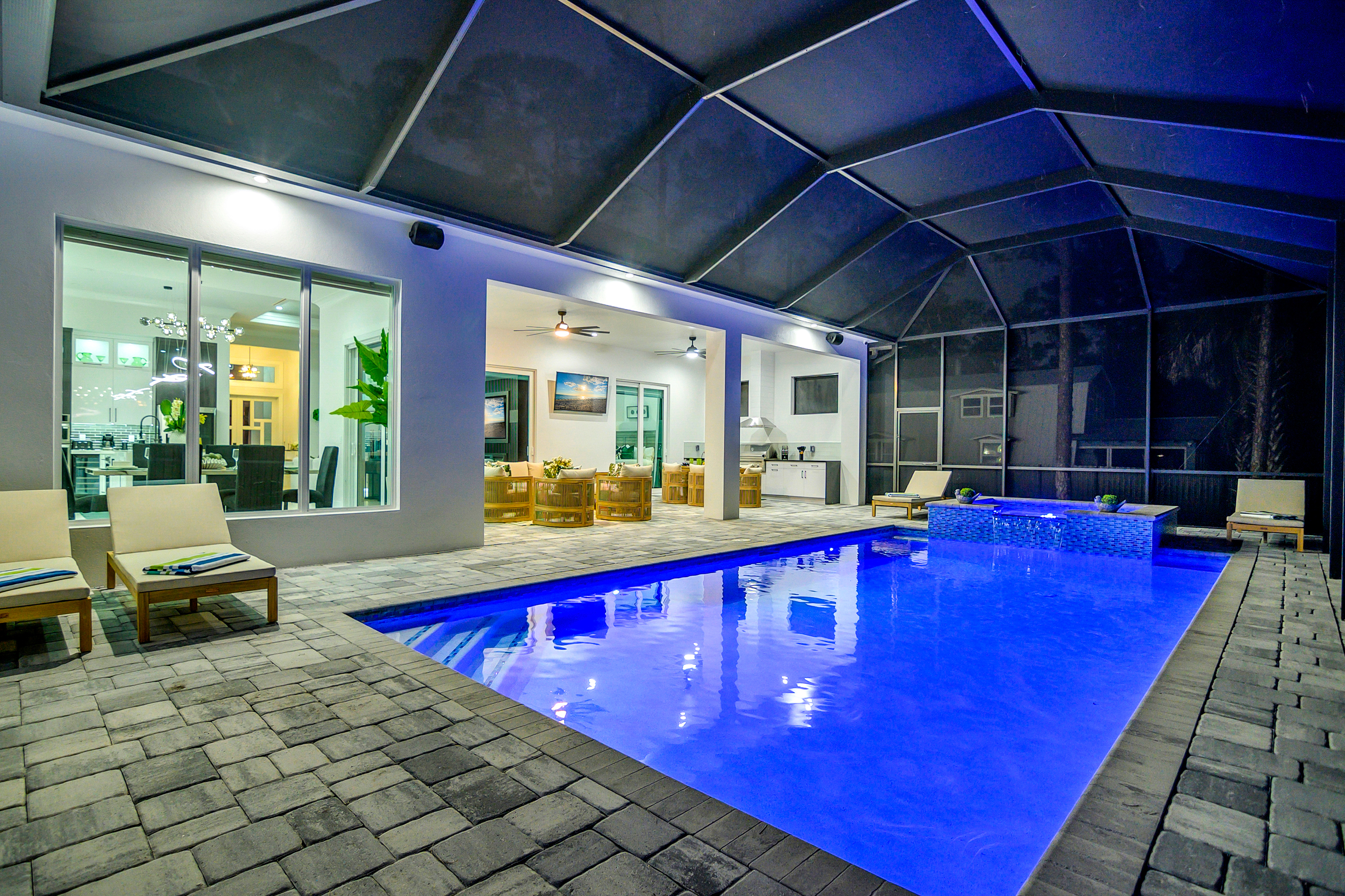 Heated pool with spa and two lounge chairs on stone patio inside screened enclosure at night