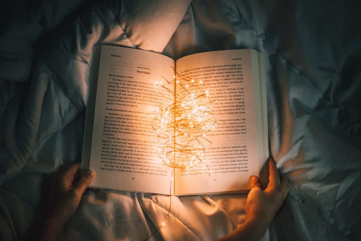 A person reading a physical book next to a glowing tablet