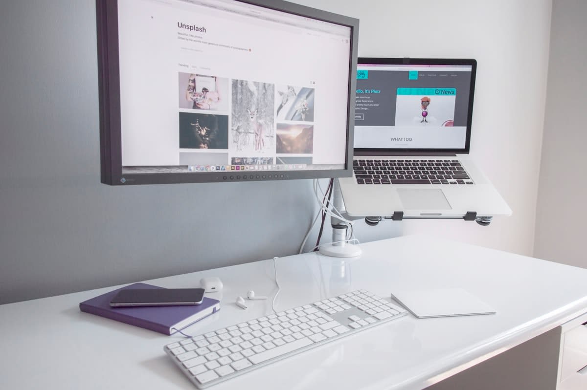 A modern minimalist workspace with multiple screens showing AI prompt chains and a physical notebook