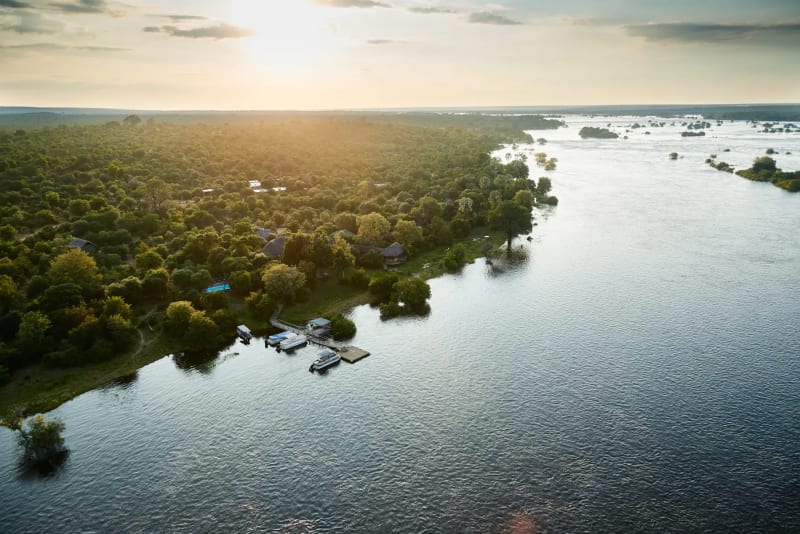 Victoria Falls River Lodge nestled on the edge of the Zambezi River with lush green trees and calm water reflecting the sky