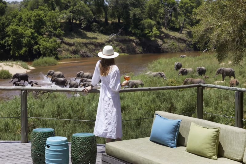 Woman wearing a sun hat standing near an outdoor lounge area with colorful cushions and a pool in a garden setting