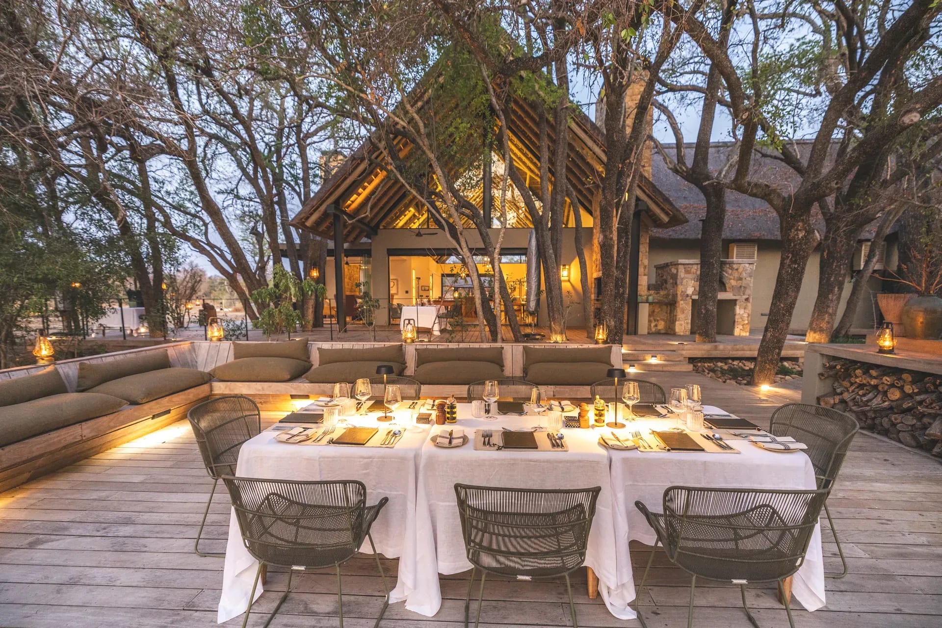 Wooden deck at Monwana Lodge with a set dining table at dusk, surrounded by trees and the lodge visible in the background.
