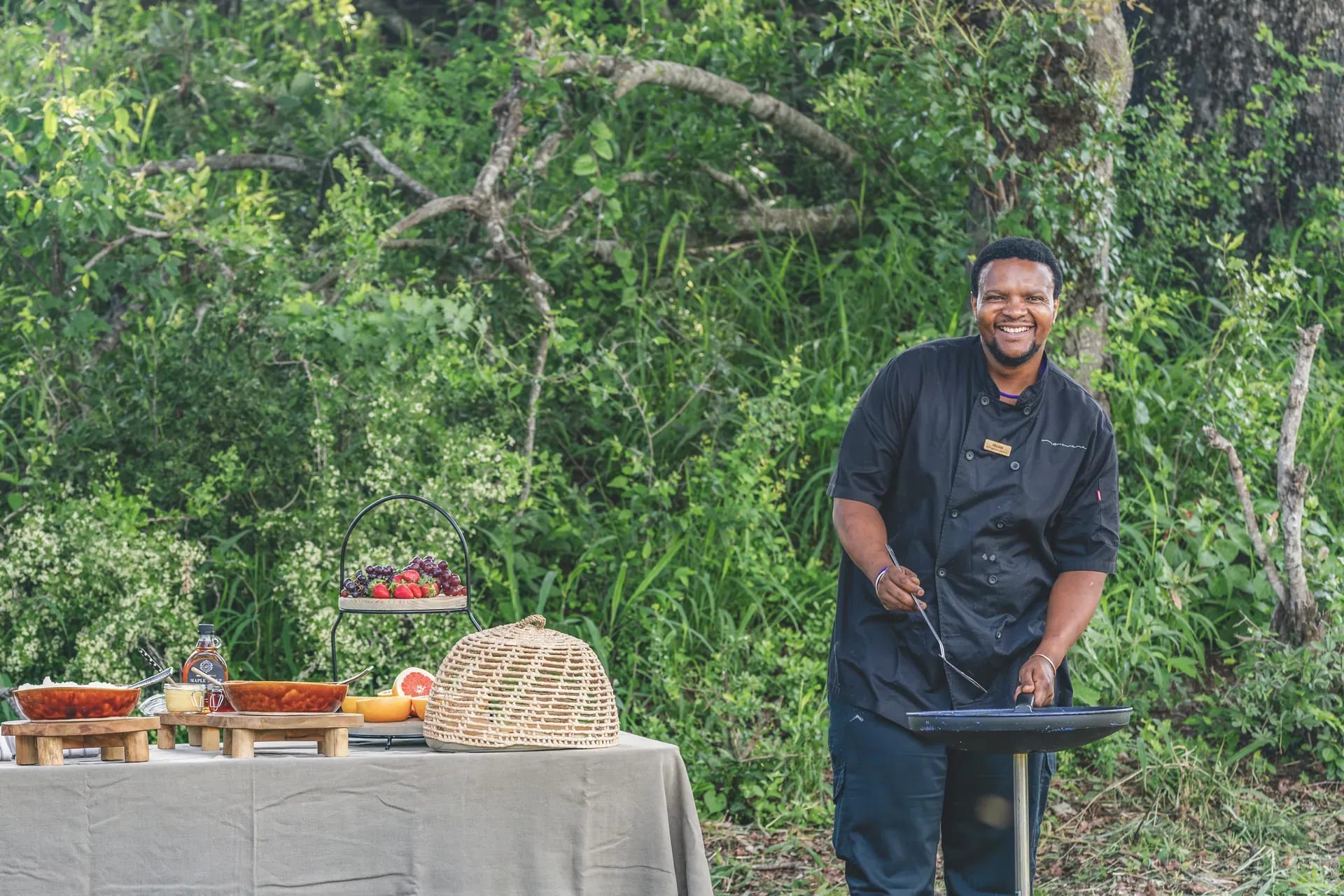 Chef preparing a meal in the African bush at Monwana Lodge, with a table laden with dishes and the bush as a natural backdrop.