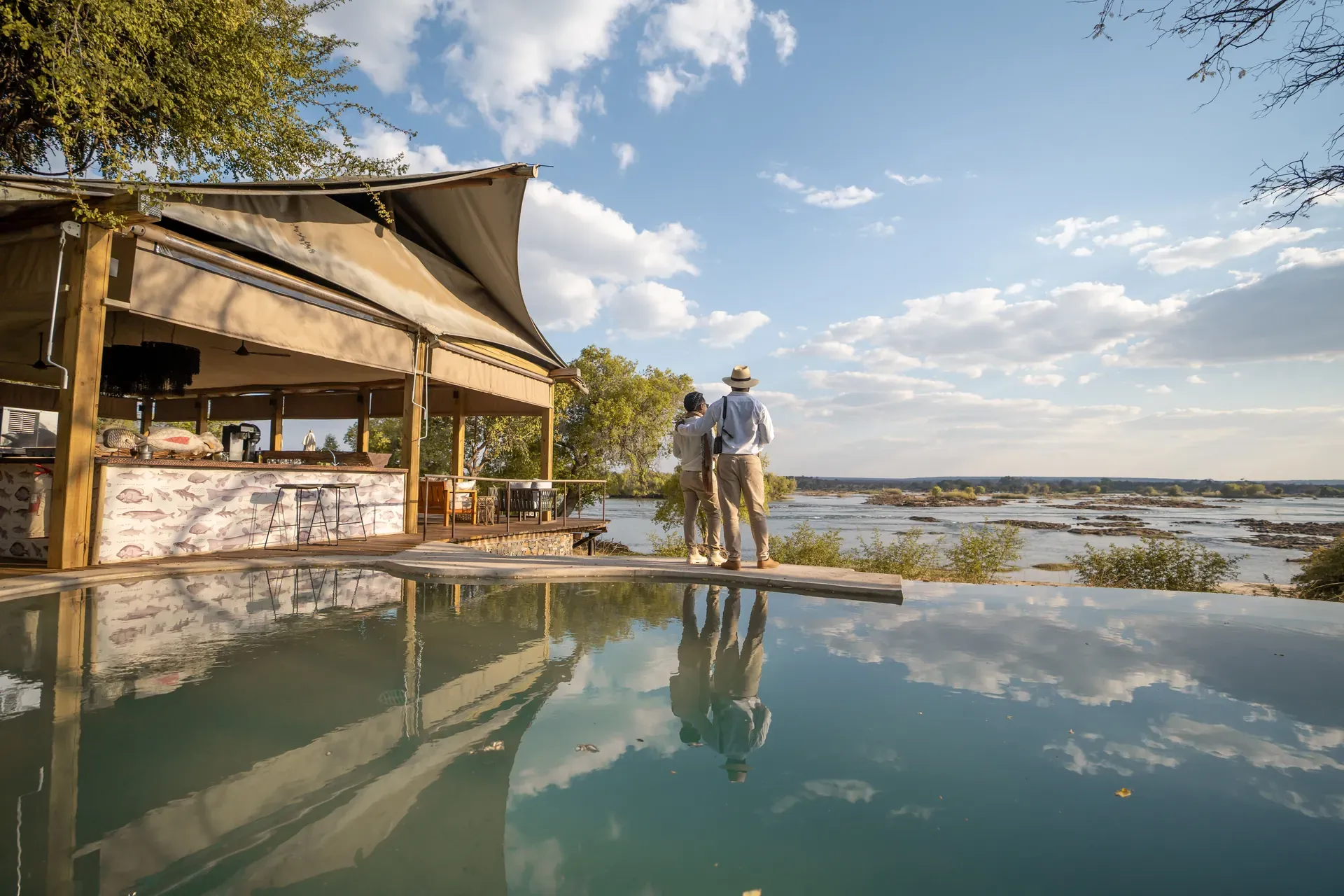 Toka Leya Camp along a tranquil river with wooden decked tents and reflections on the water under a blue sky
