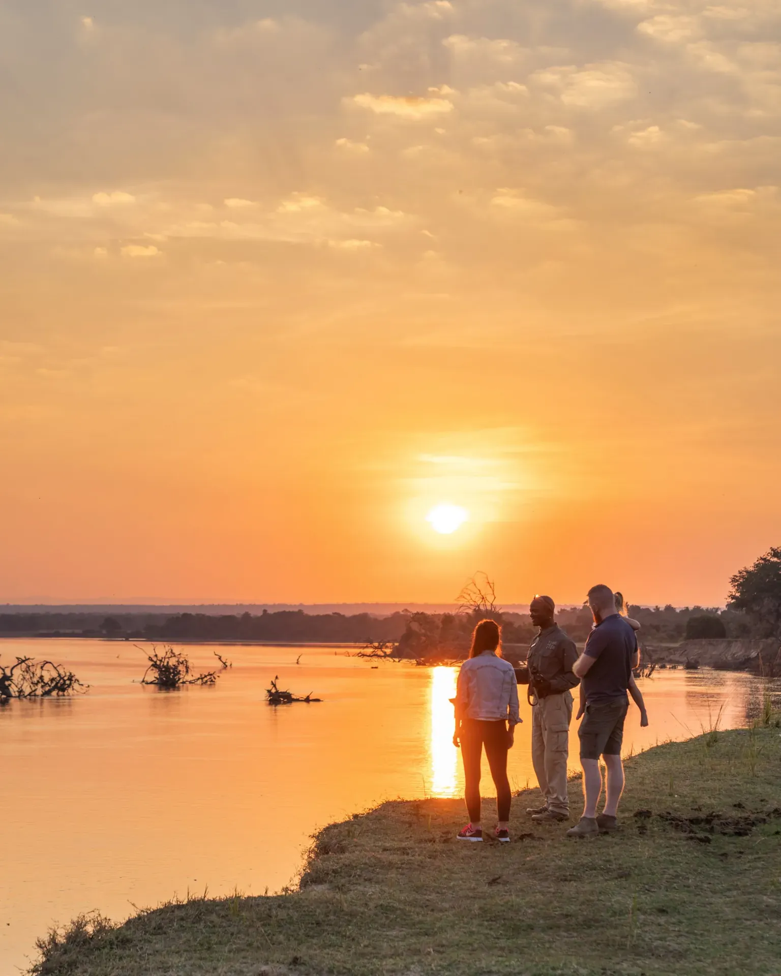 Group of people on a walking safari along a riverbank trail at sunset with a golden sky