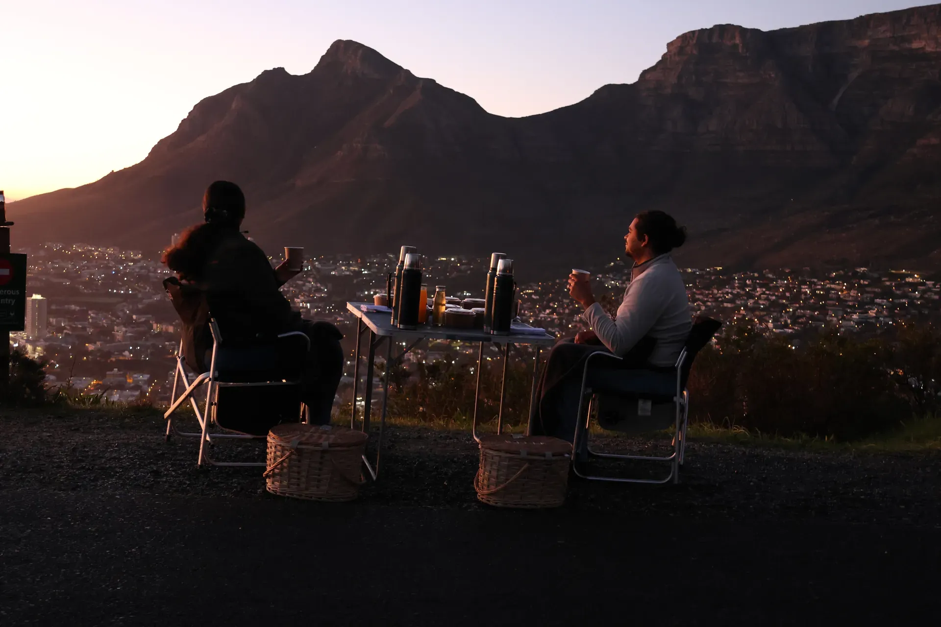 Silhouetted people gathered at dusk under string lights with mountains in the background and a campfire glow