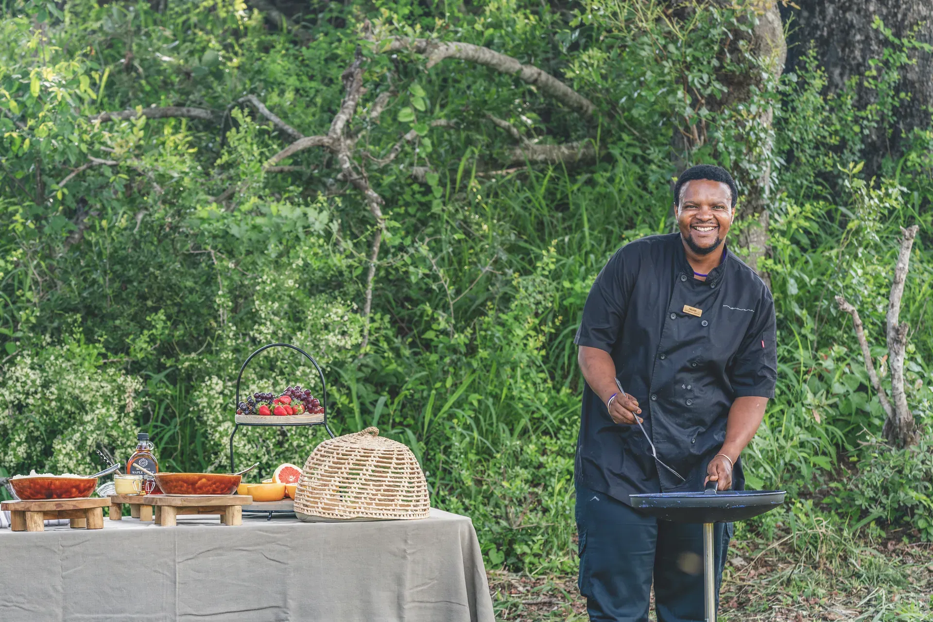 Chef preparing a meal in the African bush at Monwana Lodge, with a table laden with dishes and the bush as a natural backdrop.