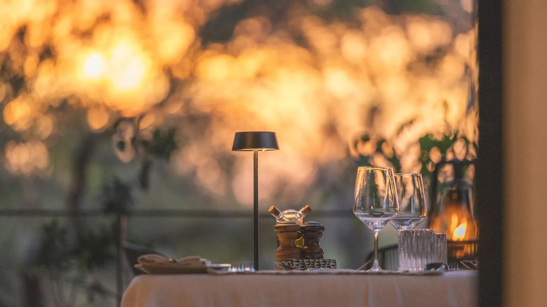 Dining table at Monwana Lodge with wine glasses and a lamp, set against a golden African bush sunset.