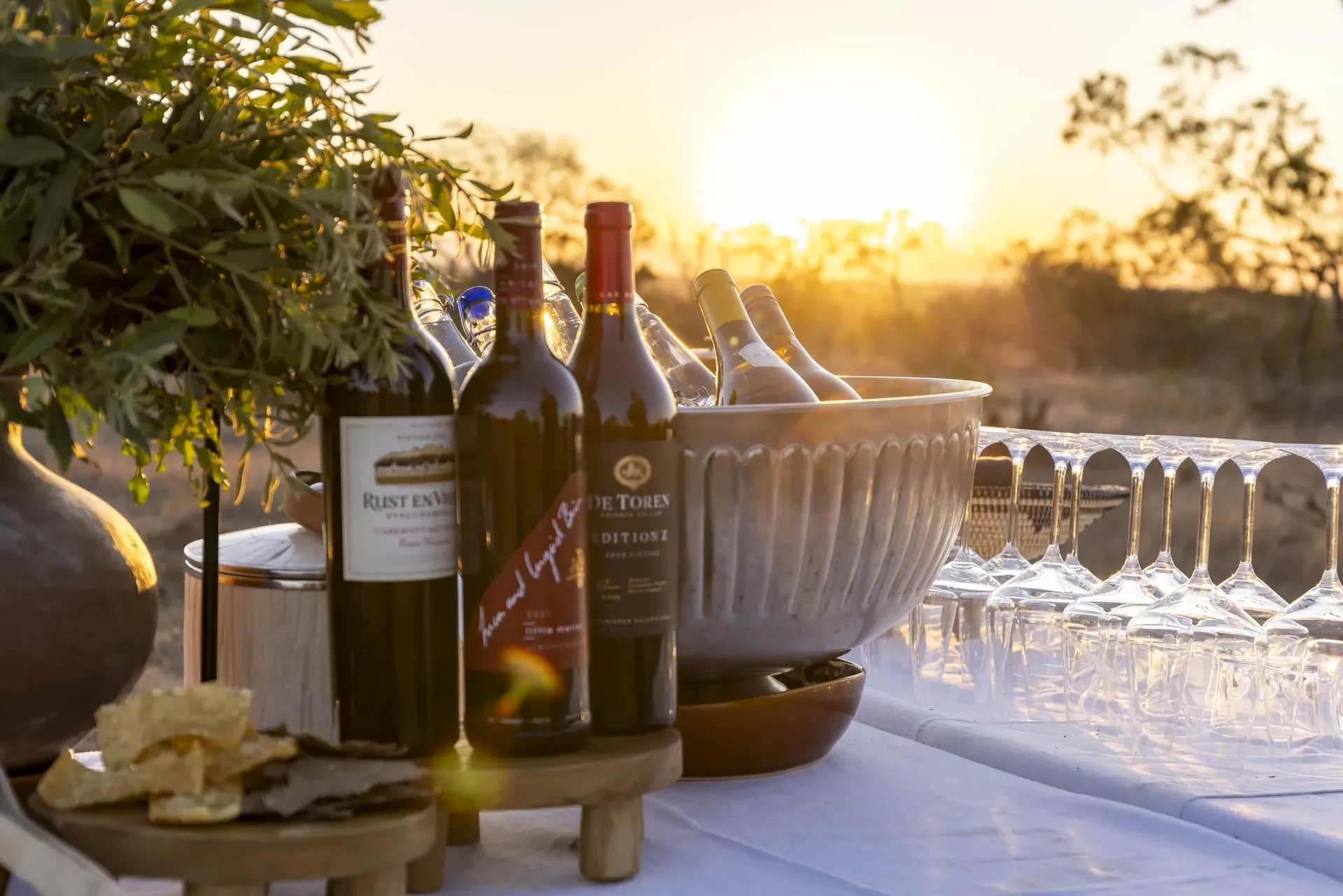 Table set with wine and glasses in the African bush at Monwana Lodge, with a sunset in the background.