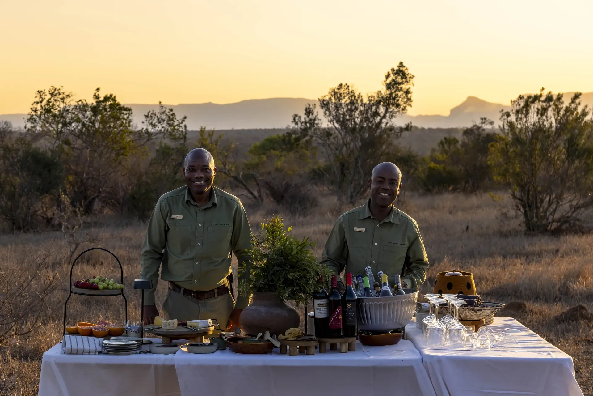 Table set with food and wine in the African bush at Monwana Lodge, with two guides standing behind it.