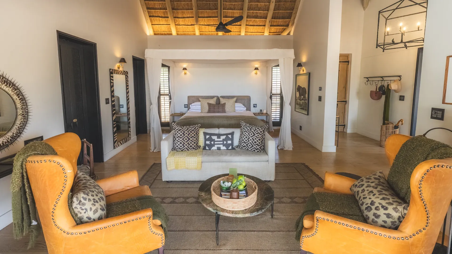 A luxurious safari-style bedroom with a high thatched ceiling, a four-poster bed draped in white fabric, and warm earthy tones.
