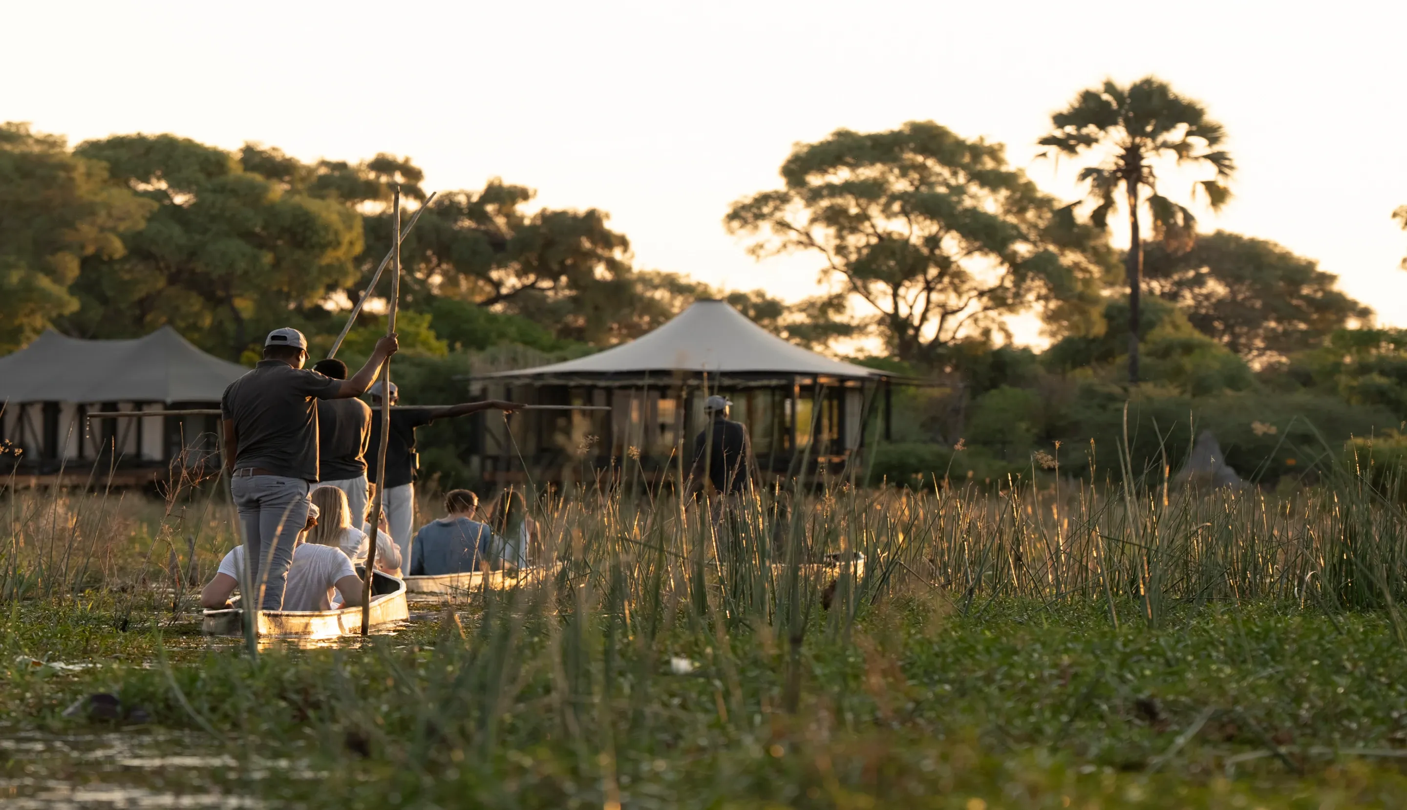 Guests in a mokoro canoe on Okavango Delta waters with Daunara Safari Camp lodge visible in the background.