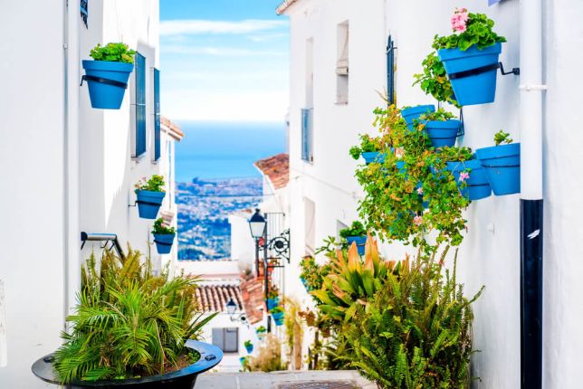 Picture shows a sunny street in the Costa del Sol, Spain. The houses are a bright white, with bright blue flowerpots.