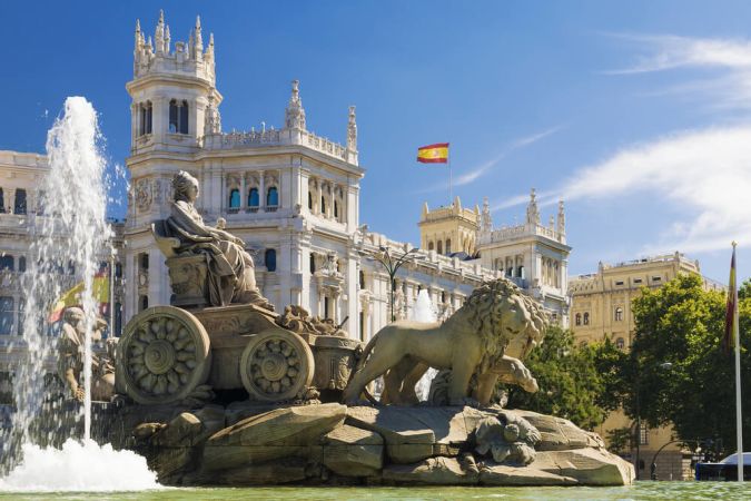 Picture shows a bright sunny day in Madrid Spain. A fountain shoots a jet of water up into the blue sky in the foreground, while majestic statues and buildings fill the background.