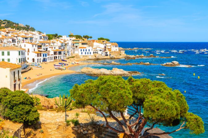 The picture shows the beautiful Spanish village of Calella de Palafrugell. A golden beach extends from the village into the bright blue sea.