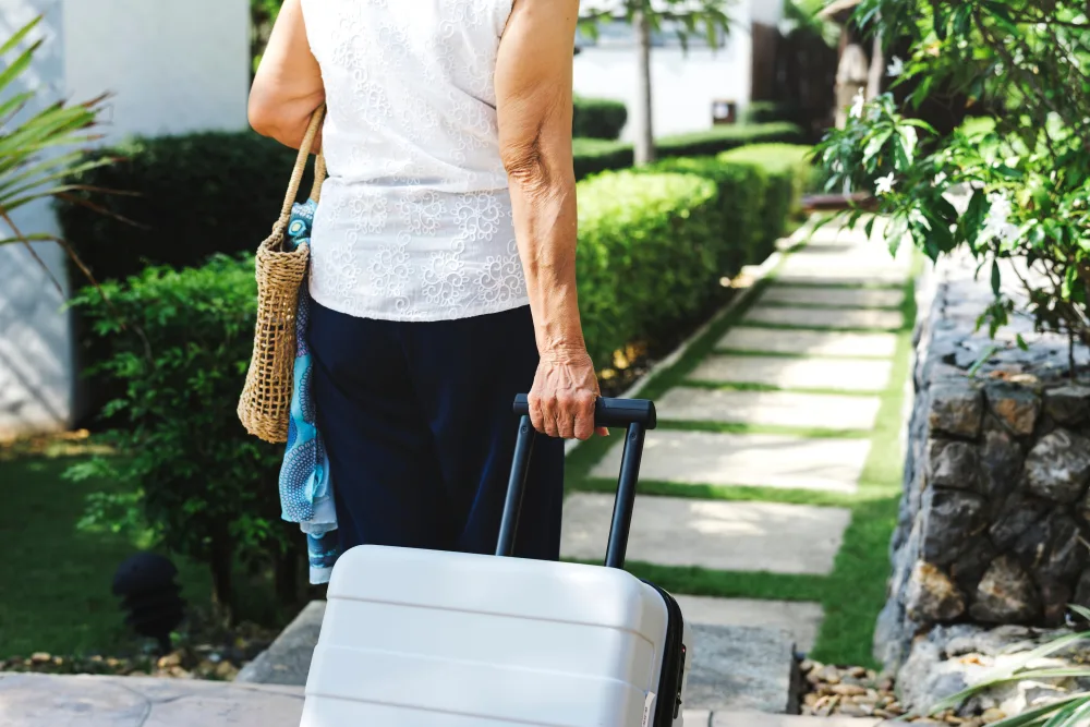 Senior woman with suitcase and beach bag walking through resort garden path, representing safe and prepared travel guided by FCDO advice.