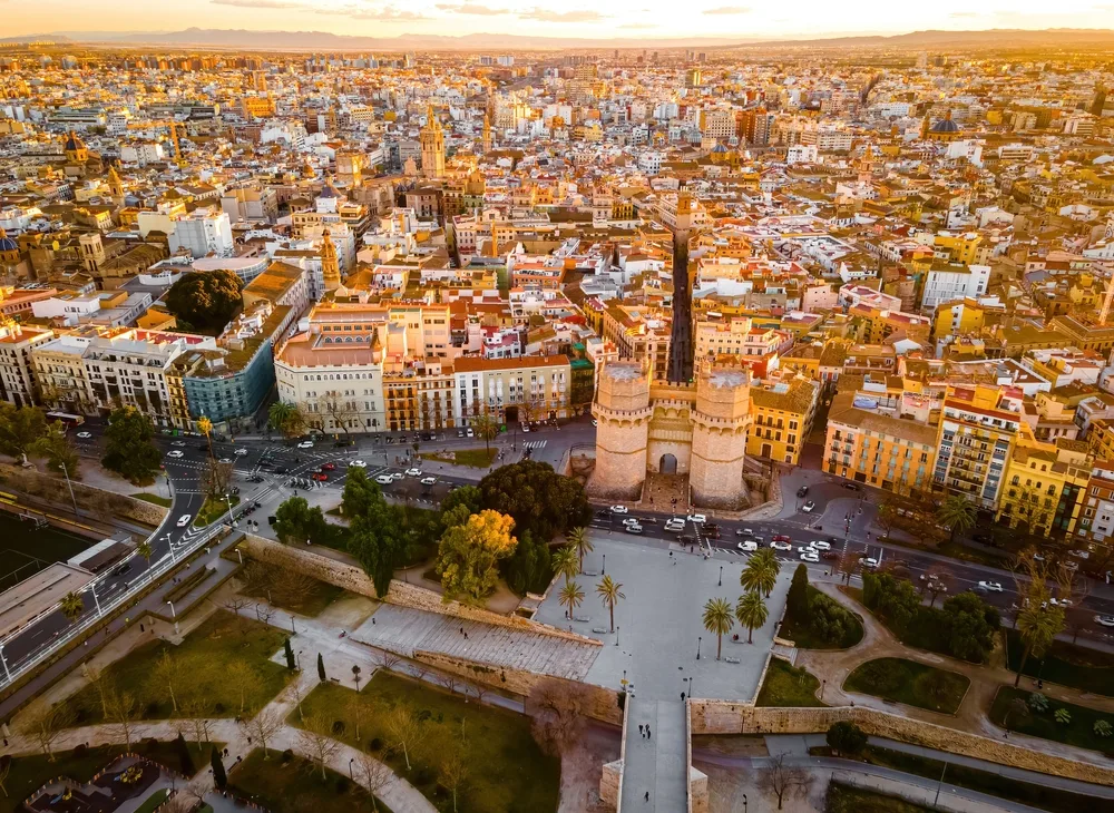 Aerial view of Valencia, Spain, featuring the historic Torres de Serranos gates, the old town’s terracotta rooftops, palm-lined gardens and city skyline at sunset.