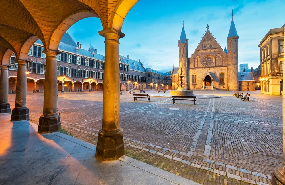 Historic Binnenhof courtyard in The Hague, Netherlands, featuring the Gothic Ridderzaal (Hall of Knights), arched colonnades, cobblestone square and benches at dusk.