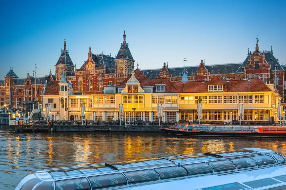 Amsterdam Central Station reflected in the canal at dusk, with historic red-brick architecture, waterfront buildings and a glass-roofed canal boat in the foreground.