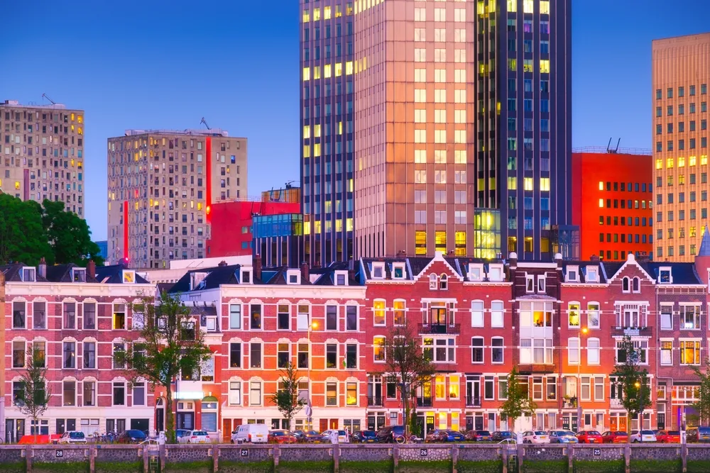 Modern Rotterdam skyline at dusk, featuring colourful high-rise buildings behind traditional Dutch townhouses along the waterfront.