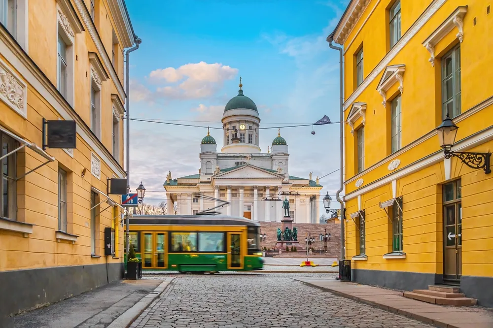 Helsinki Cathedral at Senate Square, Finland, viewed from a cobblestone street with yellow buildings and a green tram passing in the foreground.