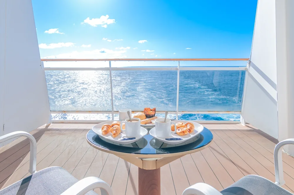 Ocean-view cruise ship balcony with breakfast table, coffee cups, and croissants under a sunny blue sky.