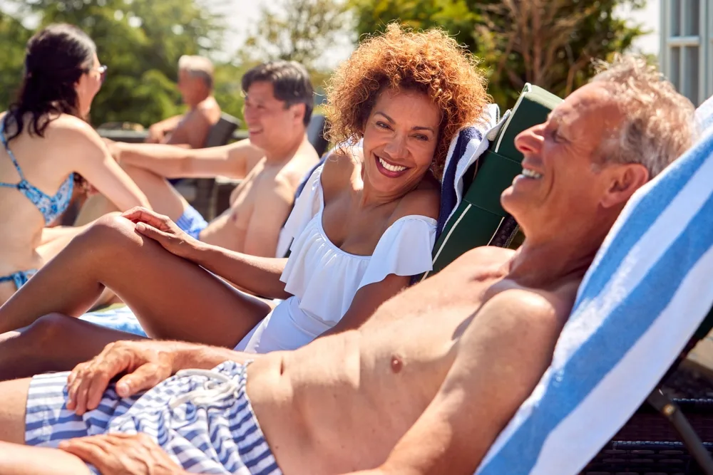 Happy couple over 50 relaxing on sun loungers by a pool, enjoying a warm holiday with friends in the background