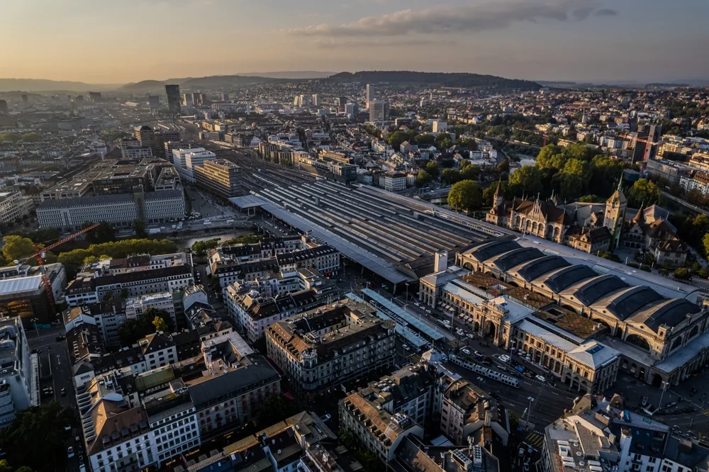 Aerial view of Zürich Hauptbahnhof train station in Switzerland, showing expansive rail platforms, historic station architecture and the surrounding city skyline at sunset