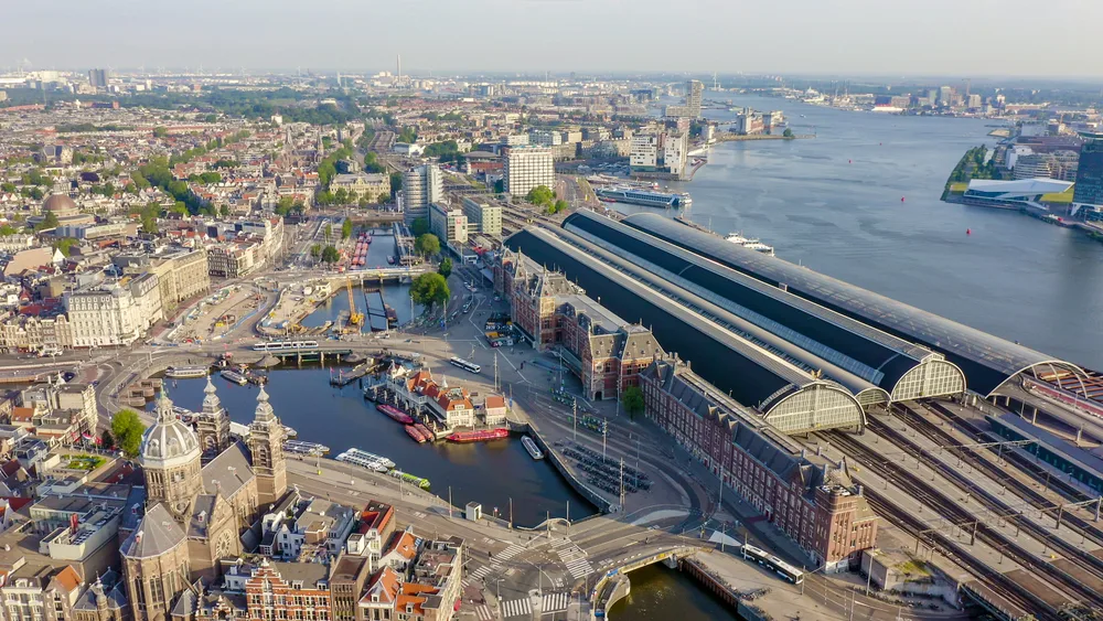Aerial view of Amsterdam Centraal Station beside canals and the IJ waterfront, with historic architecture and surrounding cityscape