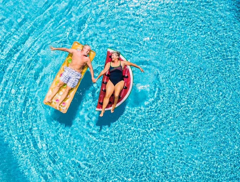 Older couple relaxing on inflatable loungers in a swimming pool, holding hands and enjoying a sunny holiday together