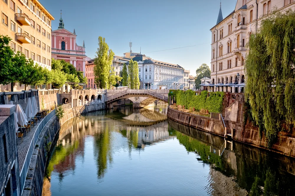 Scenic view of Ljubljana, Slovenia, featuring the Ljubljanica River, a stone bridge, historic city architecture, and tree-lined waterfront reflections on a clear sunny day.