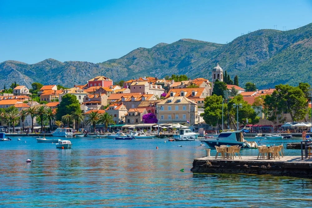 Coastal town on Croatia’s Adriatic coast with orange-roofed buildings, a waterfront marina, boats in a turquoise harbour, and rugged mountains rising in the background under a clear blue sky.