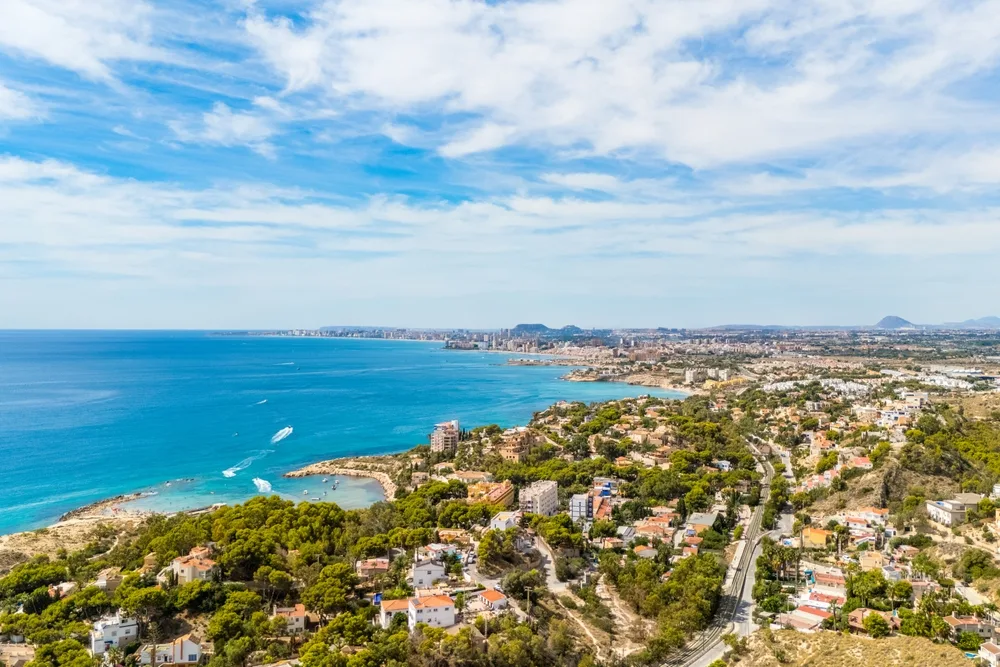 Aerial view of Alicante’s Mediterranean coastline in Spain, featuring turquoise waters, sandy coves, coastal neighbourhoods, and a sprawling seaside city beneath a bright blue sky.