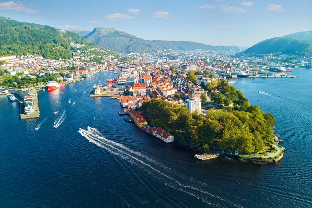 Aerial view of Bergen, Norway, with colourful waterfront buildings, boats on the harbour and green mountains surrounding the city on a sunny day.
