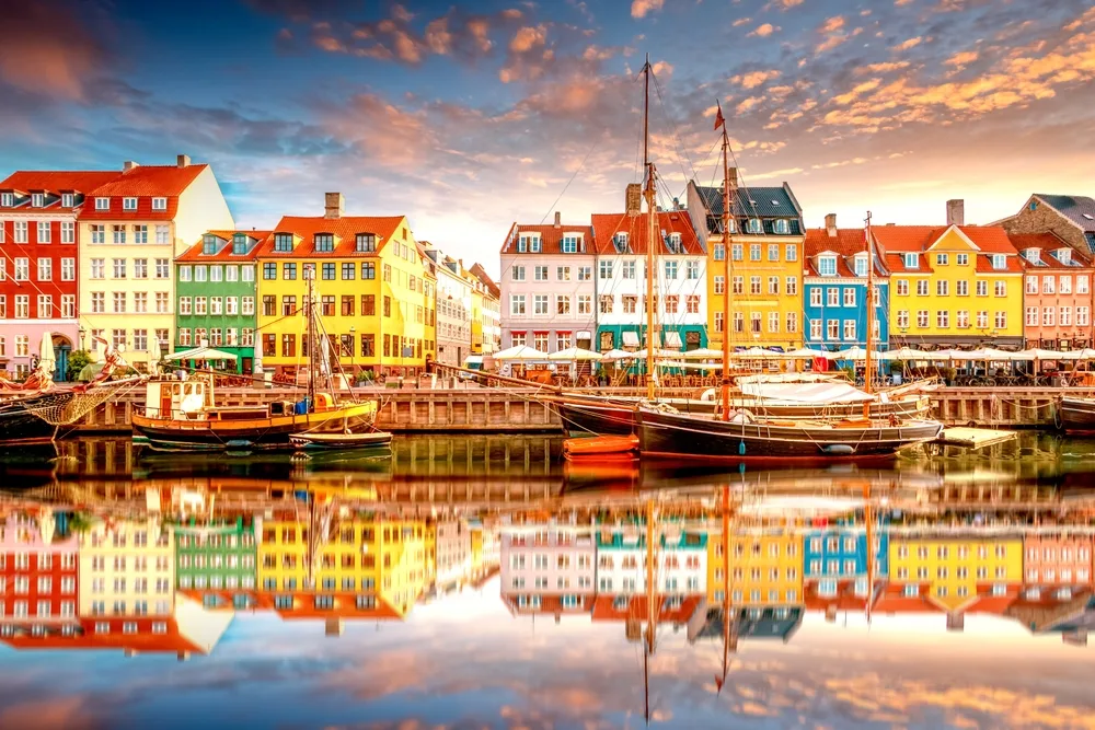 Colourful waterfront buildings and traditional sailing boats at Nyhavn Harbour in Copenhagen, Denmark, reflected in calm water at sunset.