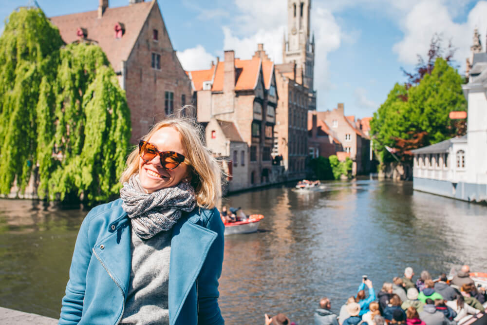 Picture shows a smiling woman enjoying Bruges in the sun. Behind her is one of the cities canals, with boats full of sightseers. Beyond the canal are the many historic buildings the city is famous for.