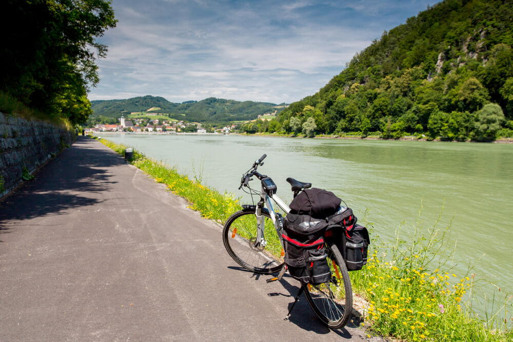 The picture shows a bike one the Danube cycle path, next to a wide river with heavily forested hills. In the distance a quaint town.