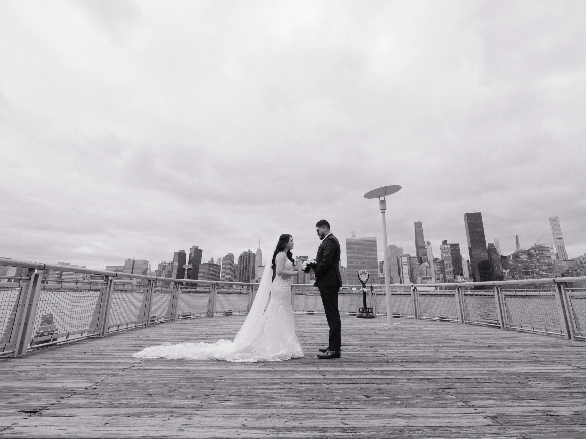 Ultra-wide first-look on a pier by the East River — the two of them small against open water.