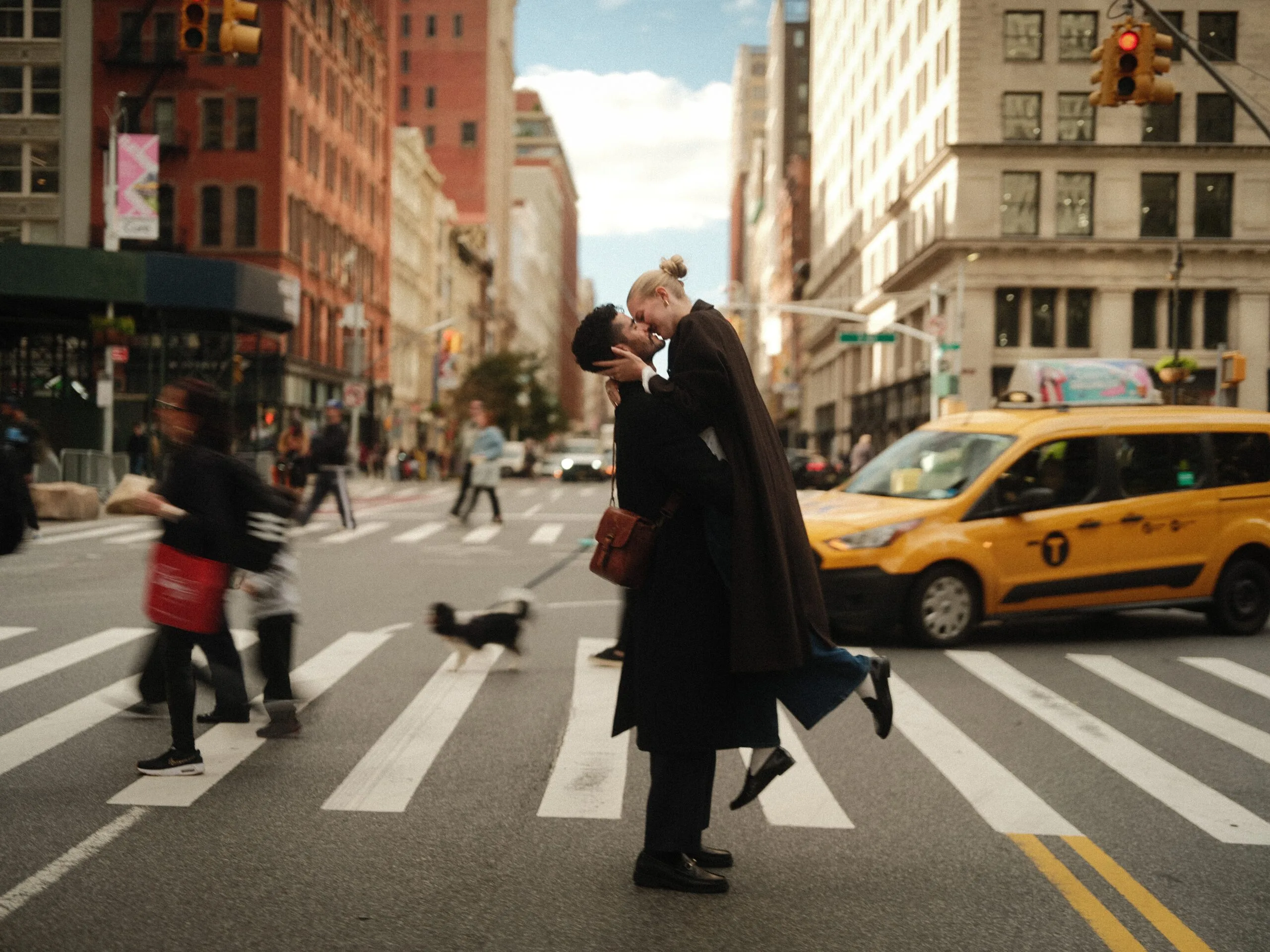 A couple lifted in a kiss mid-crosswalk on Fifth Avenue, Flatiron behind, pedestrians blurring past.