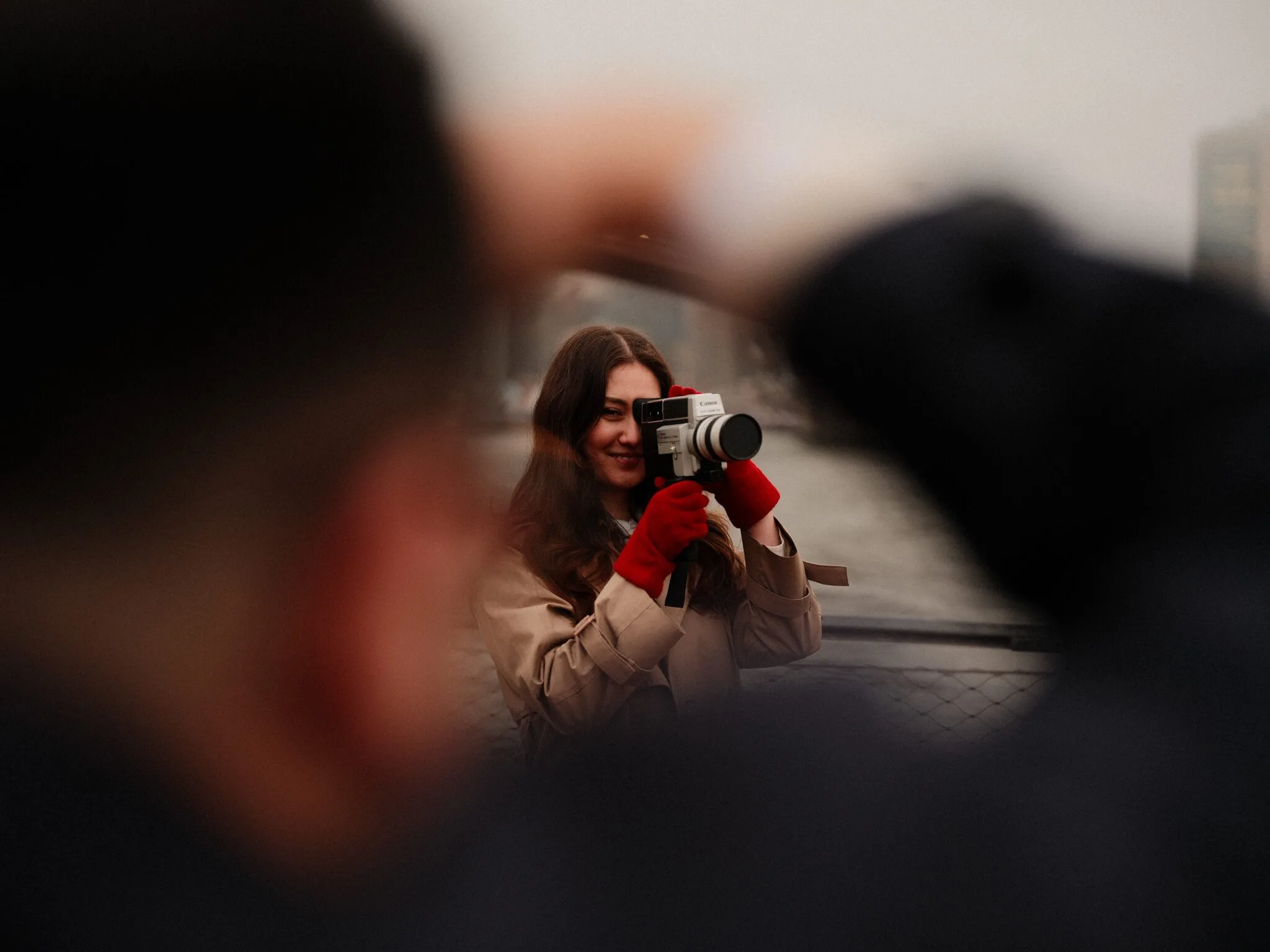 Over Alfredo's shoulder — Lexi lifts the Super-8 to her eye, smiling wide, the focus tight on her face with him blurred in the foreground.