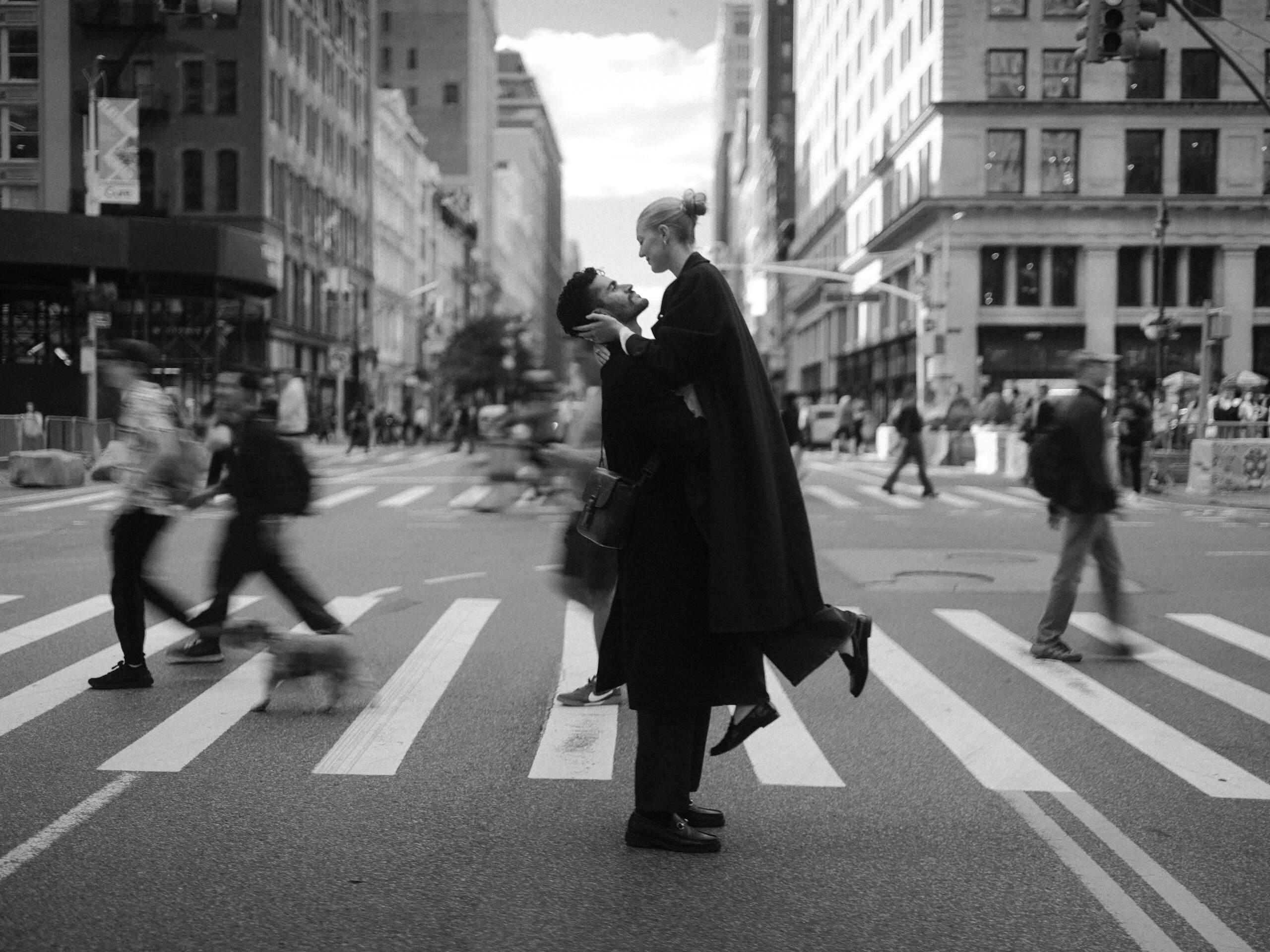Couple embracing in the middle of a New York City crosswalk, black and white, pedestrians blurred around them